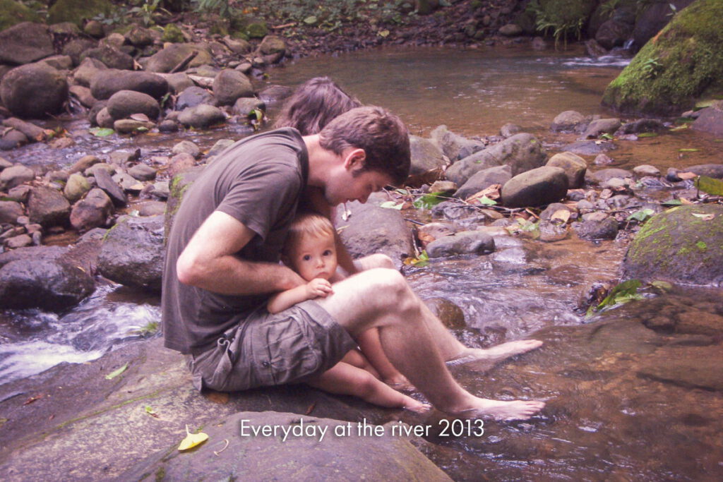 A father and a small child sitting together in a shallow river among boulders