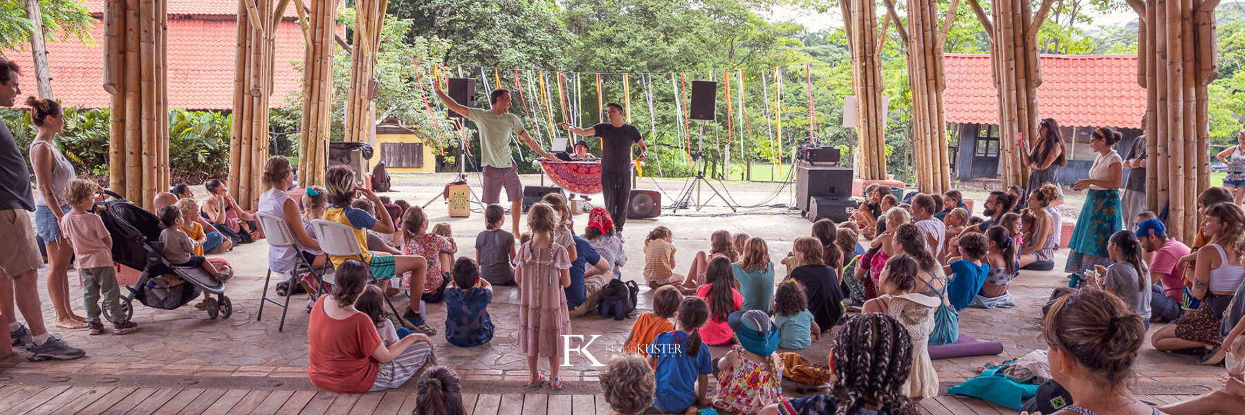 Families and children gathered in a circle under an open bamboo pavilion at a Machuca Valley community event